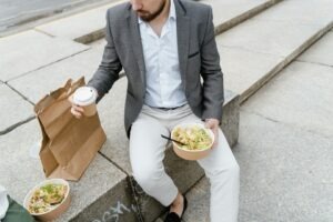 Un bărbat este așezat pe o bordură, mănâncă o salată și bea o cafea